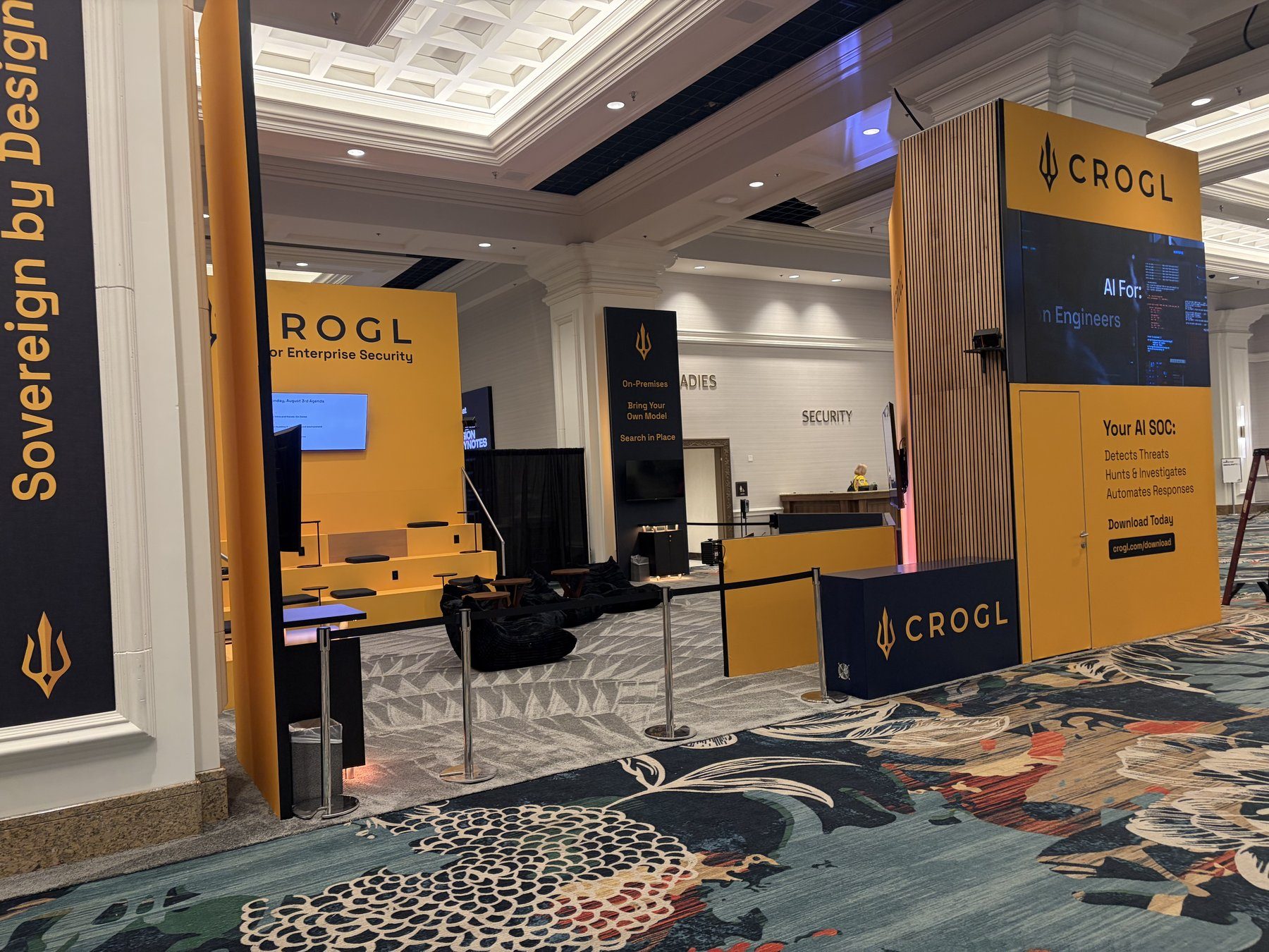 Wide view of the full Crogl booth footprint: a Sovereign by Design banner, the orange lounge with tiered seating, and the video tower