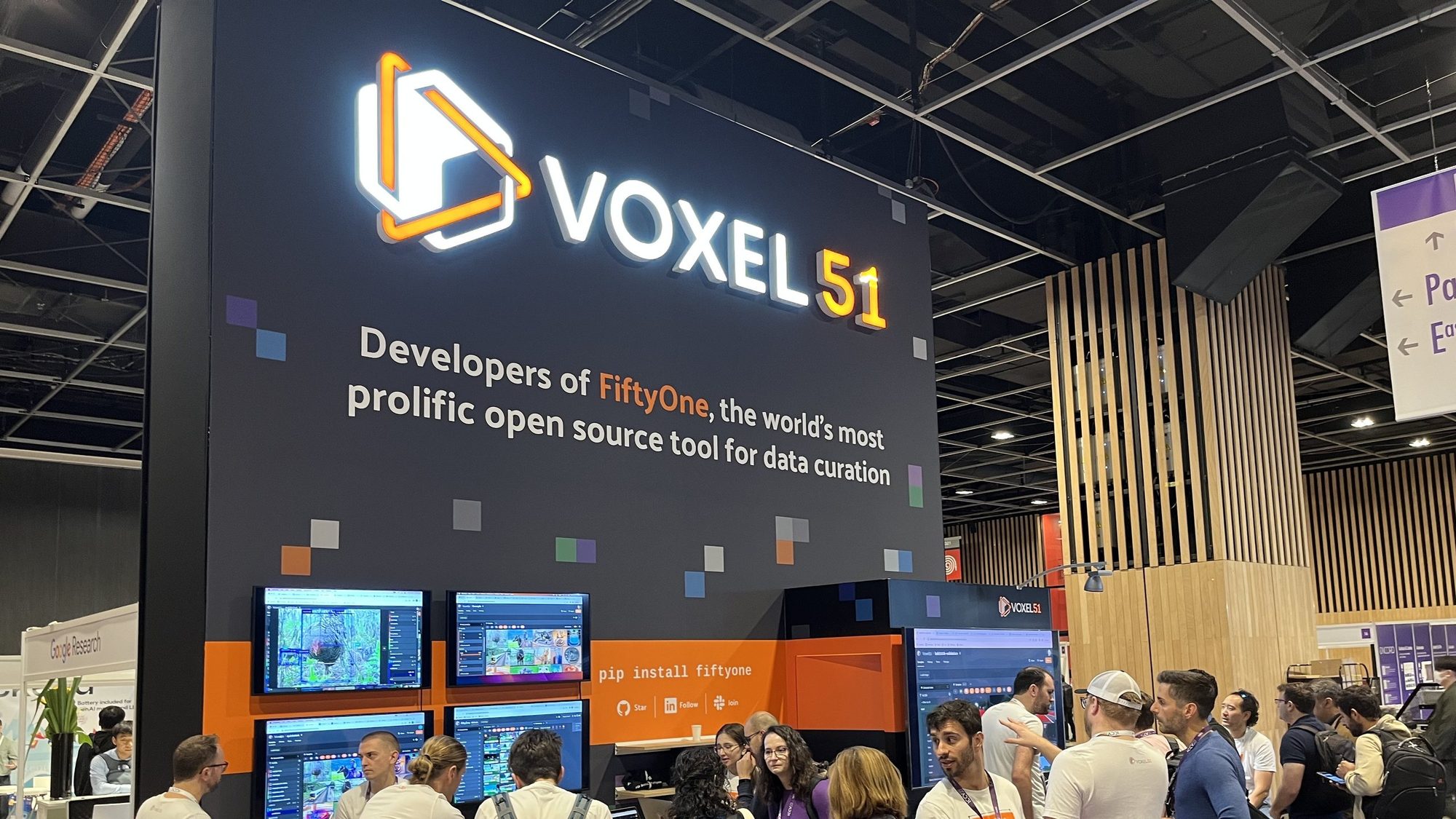 Voxel51 conference booth with the lit logo sign, the FiftyOne headline wall, and a crowd of developers at the demo screens